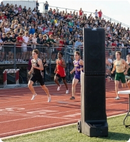 Bigfoot 3 portable line array speaker system positioned trackside at a high school athletics event, providing clear announcements and audio coverage as runners compete on the track in front of a packed stadium crowd. Bigfoot 3 portable line array speaker system positioned trackside at a high school athletics event, providing clear announcements and audio coverage as runners compete on the track in front of a packed stadium crowd.