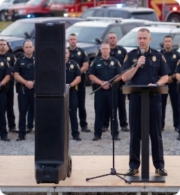 Bigfoot 3 portable line array speaker system used at a public safety event, standing beside a law enforcement officer delivering remarks to a crowd, with first responders gathered in the background for clear speech reinforcement and community communication. Bigfoot 3 portable line array speaker system used at a public safety event, standing beside a law enforcement officer delivering remarks to a crowd, with first responders gathered in the background for clear speech reinforcement and community communication.